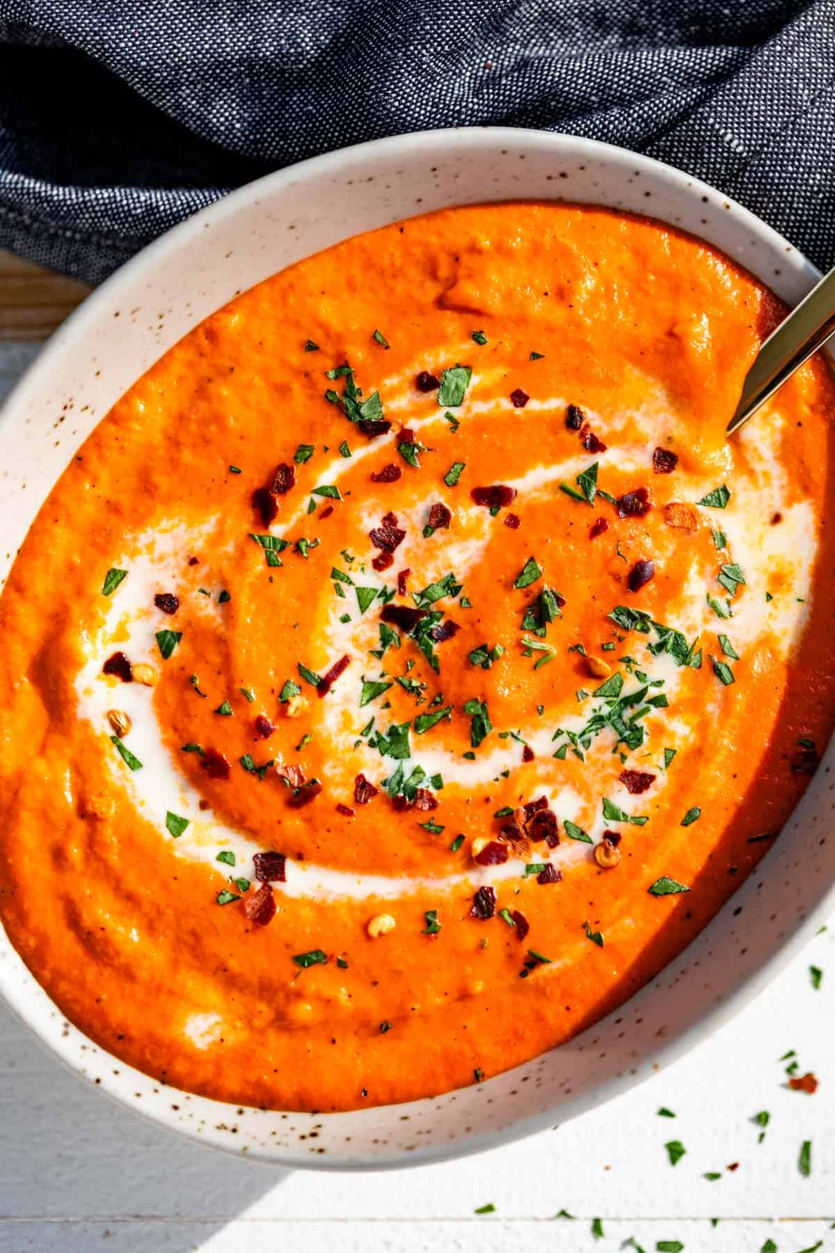 A pottery bowl filled with roasted red pepper sauce topped with a drizzle of cream, chopped parsley, and red pepper flakes.