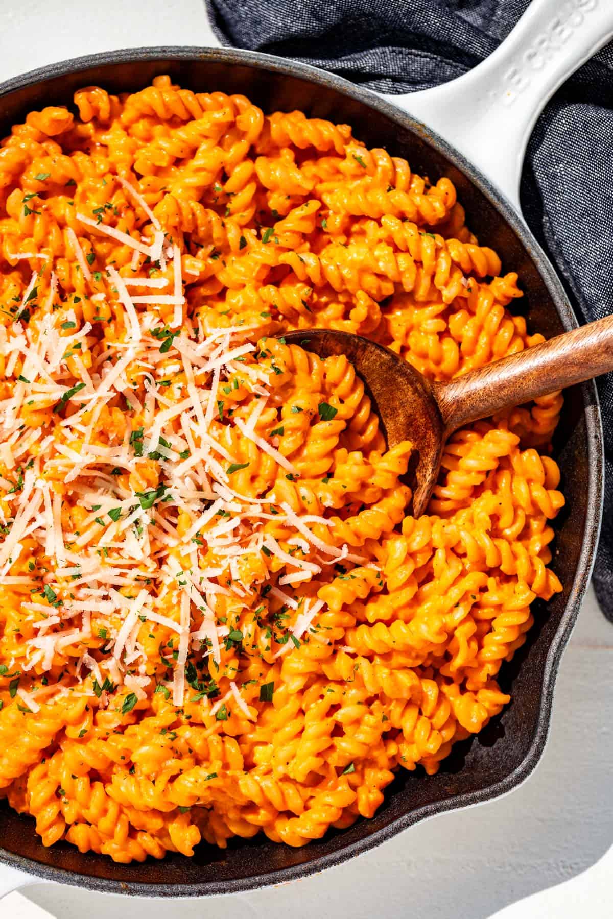 Roasted red pepper sauce tossed with fusilli pasta in a white cast iron skillet with a wood spoon in it.