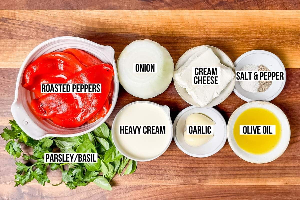 Roasted red peppers, basil, parsley, heavy cream, garlic, olive oil, cream cheese, sea salt and pepper and onion in bowls on a wood cutting board.