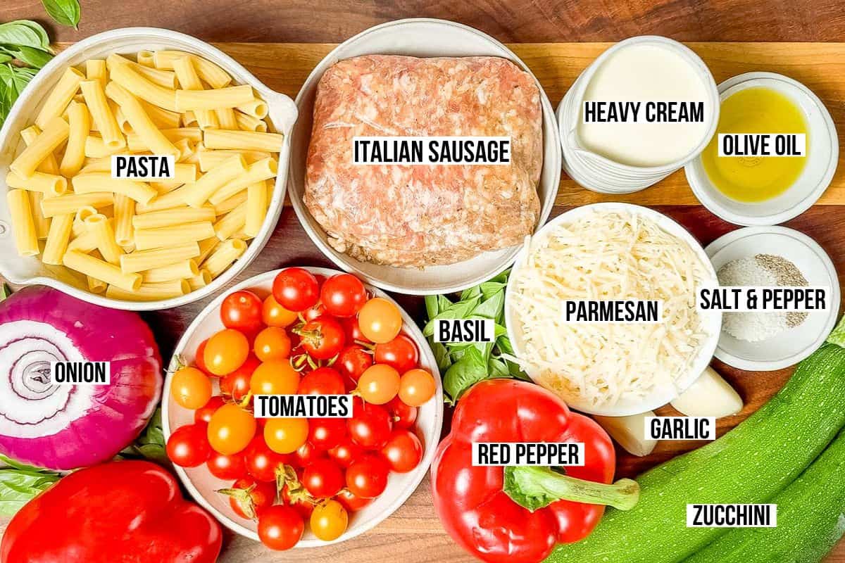 Pasta, Italian sausage, peppers, onion, parmesan cheese, garlic, basil, heavy cream. cherry tomatoes, zucchini and olive oil in pottery bowls on a wood cutting board.