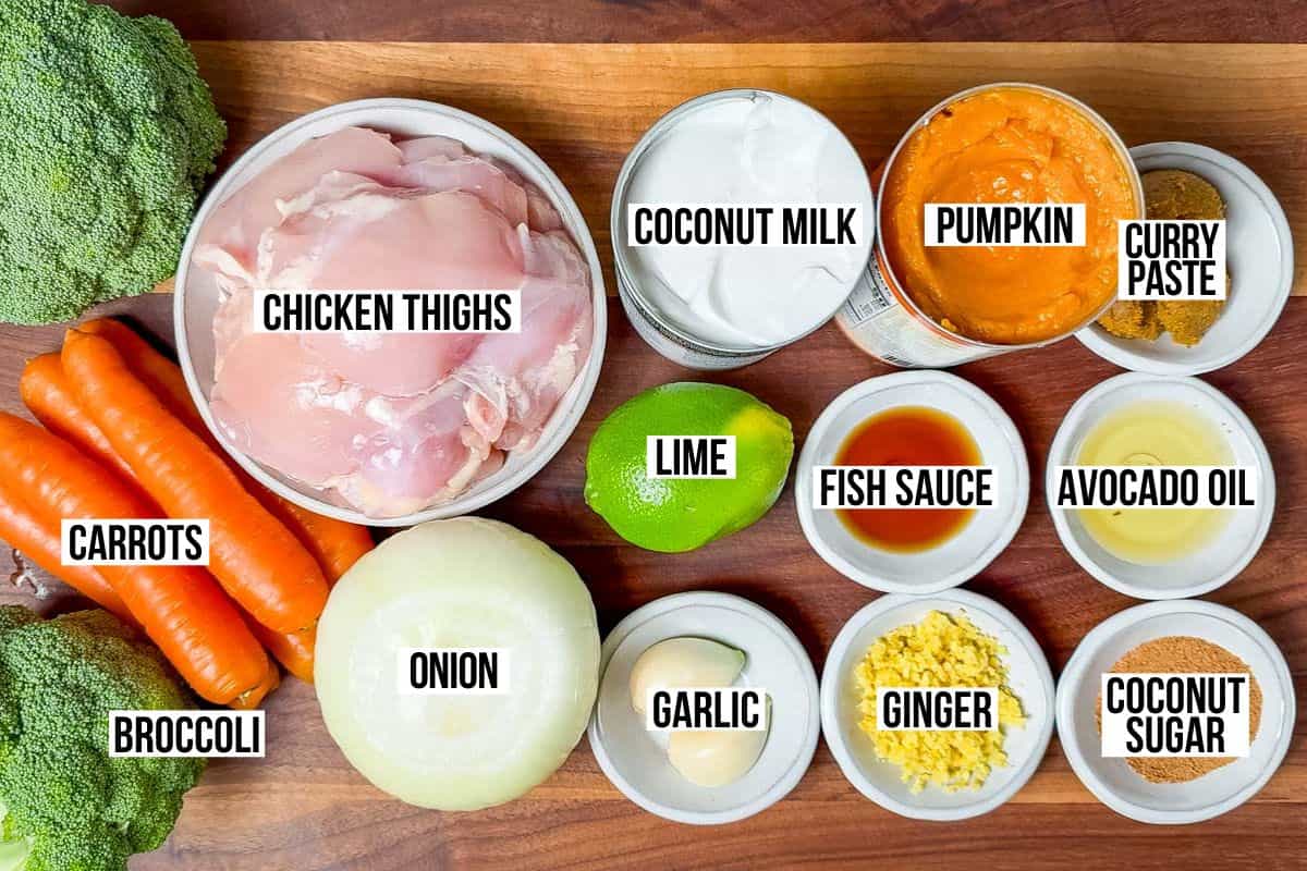 Chicken thighs, curry paste, garlic, ginger, pumpkin purée, lime, coconut milk, carrots, broccoli, lime, and coconut sugar in bowls on a wood cutting board.