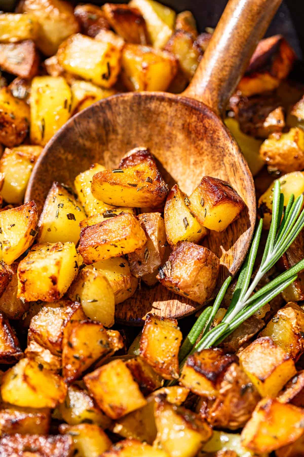 A wood spoon scooping up pan fried potatoes with a sprig of rosemary on the side.