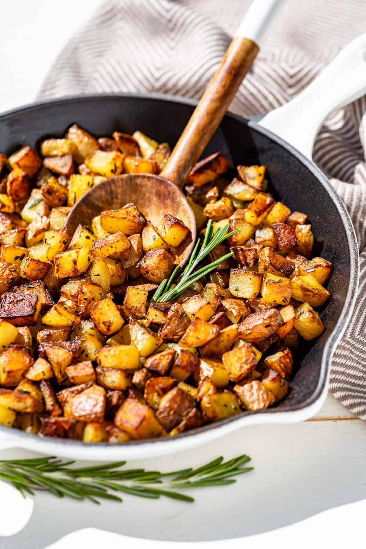 Pan fried potatoes in a white cast iron skillet with a wood spoon scooping up some potatoes and sprigs of rosemary on the side.