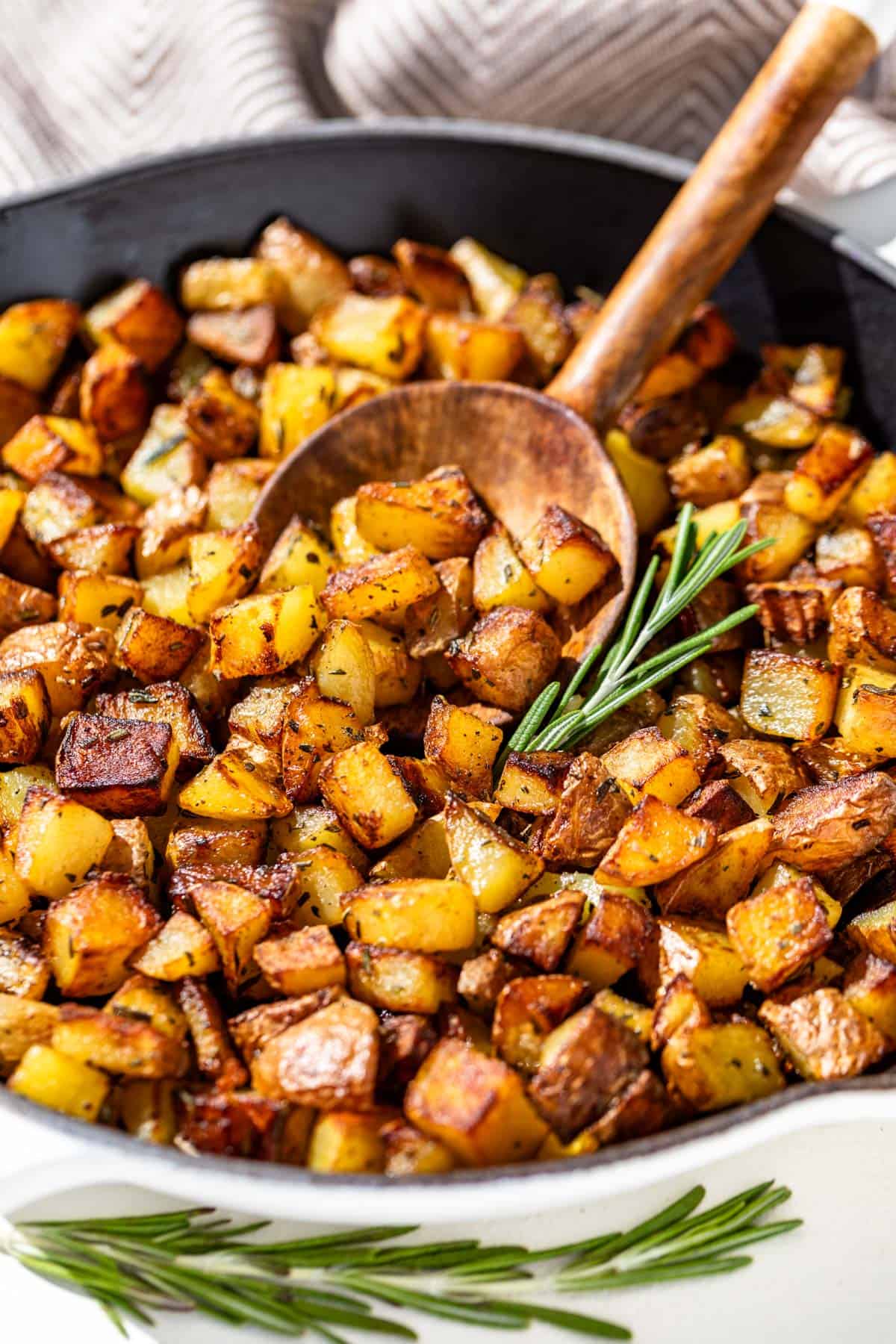 Golden brown pan fried potatoes in a skillet with a wood spoon in them with a sprig of rosemary by the pan.
