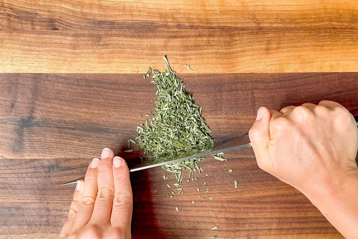 Mincing the rosemary with a chef's knife on a wood cutting board.