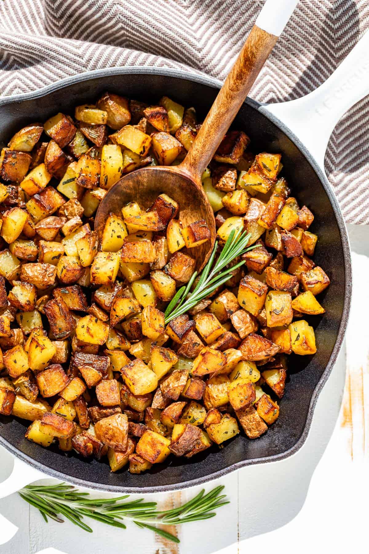 Browned pan fried potatoes in a white cast iron skillet with a wood serving spoon in it topped with a sprig of rosemary.