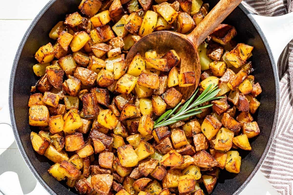 Pan fried potatoes in a white cast iron skillet with a wood spoon scooping some out.